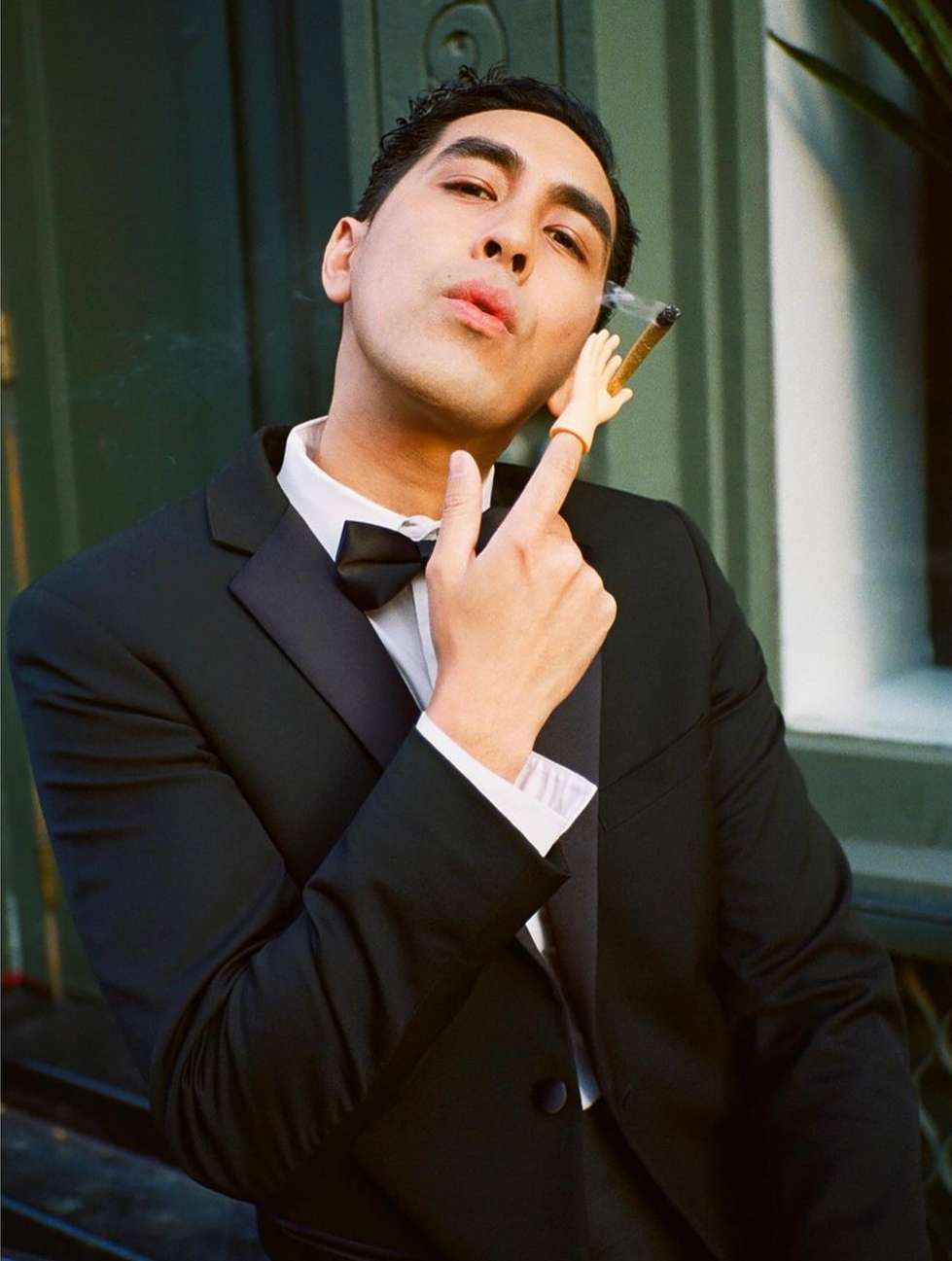 Man in tuxedo strikes pose outdoors with toy hand and cigarette, offering a playful take on formal wear and smoking.