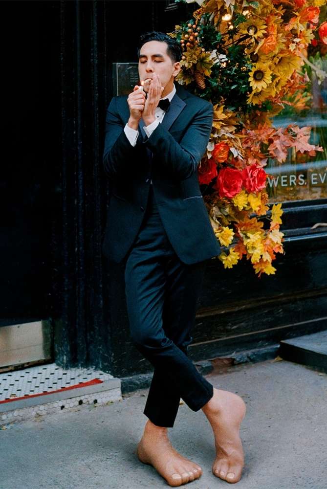 A man in a tuxedo lights a cigar while standing barefoot outside a flower shop, surrounded by autumn-toned floral arrangements.