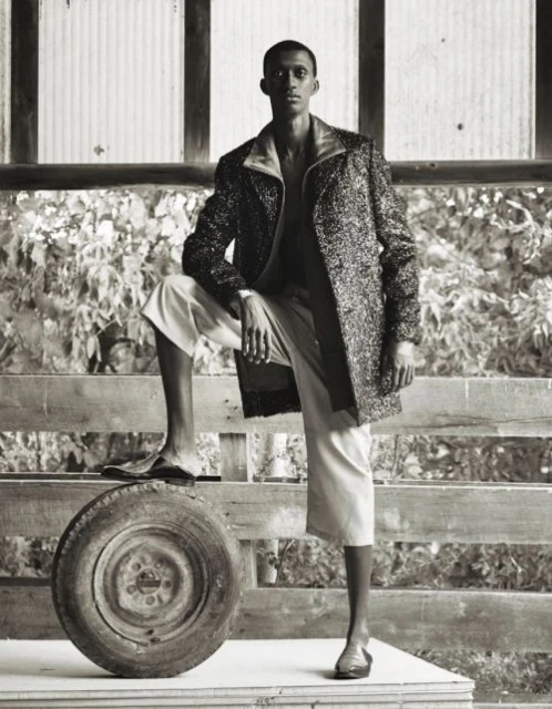 A stylish Black male model poses with a tire and fence wearing a coat, cropped pants, and sandals in a creative fashion shot.