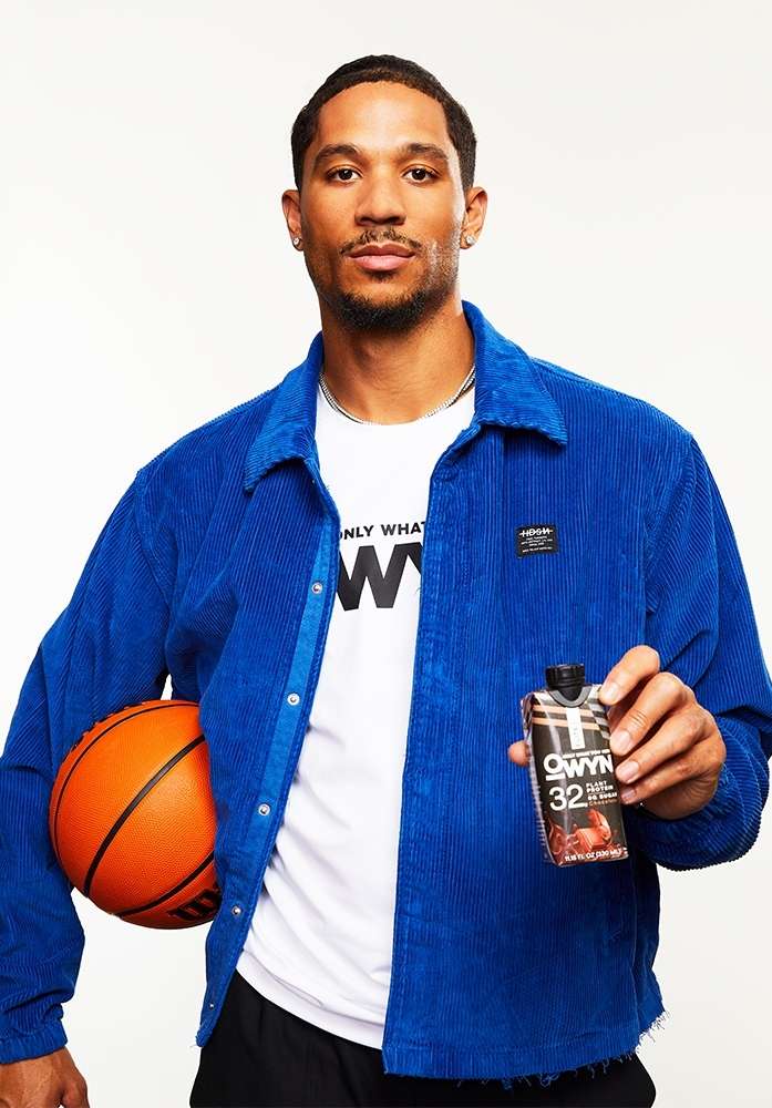 Athletic African-American man posing in a blue corduroy jacket holding a basketball and OWYN protein shake in a bright photo studio.