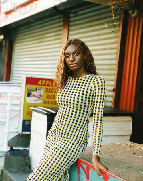 An adult female poses outside a building wearing a yellow-and-black checkered dress with long brown hair, accessorized with hoop earrings.