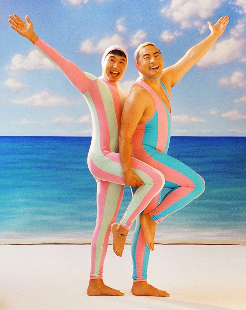 Two Asian adults in striped bodysuits stand close on a sandy beach with arms raised against a backdrop of blue sea and sky with clouds.