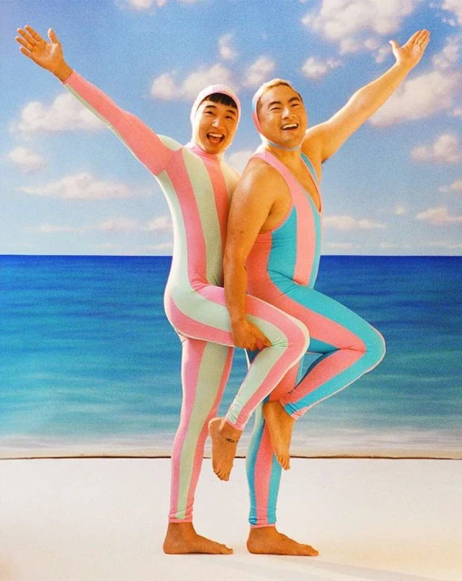 Two Asian adults in striped bodysuits stand close on a sandy beach with arms raised against a backdrop of blue sea and sky with clouds.