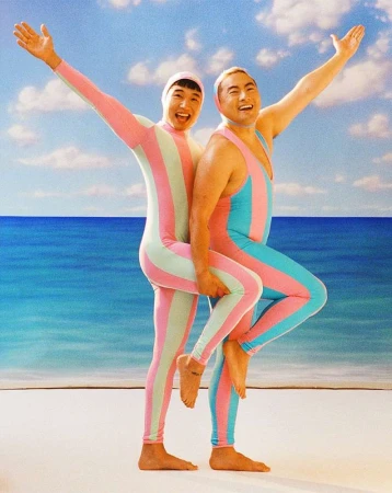 Two Asian adults in striped bodysuits stand close on a sandy beach with arms raised against a backdrop of blue sea and sky with clouds.