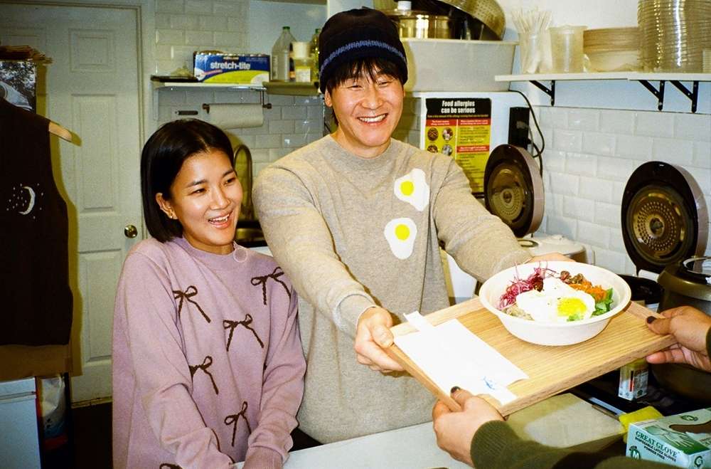 Two smiling people indoors, one holding a wooden tray with a poke bowl; food preparation and presentation in a bright kitchen.