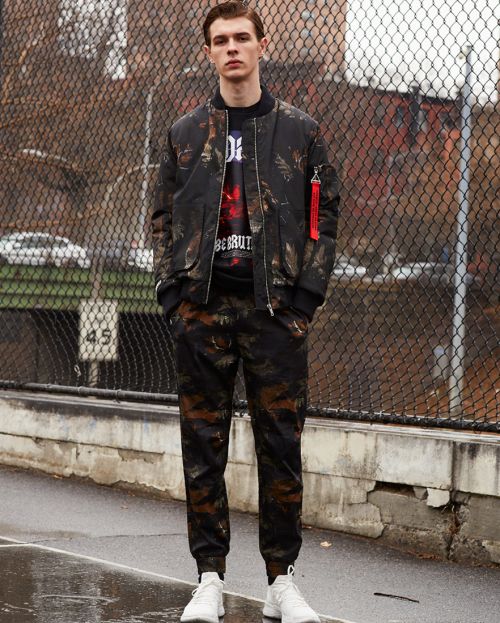A young male model in a camouflage bomber jacket and matching pants stands outdoors, showcasing a fashion-forward, urban style.