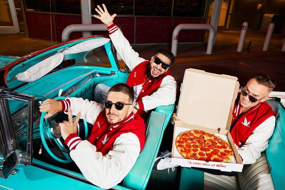 Three adult males in varsity jackets pose in a vintage turquoise convertible car with a pizza box, creating a retro vibe.