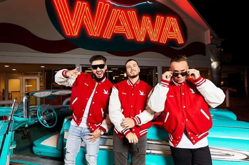 Three men pose in front of a Wawa store and a turquoise vintage car, wearing matching red and white jackets and sunglasses at night.