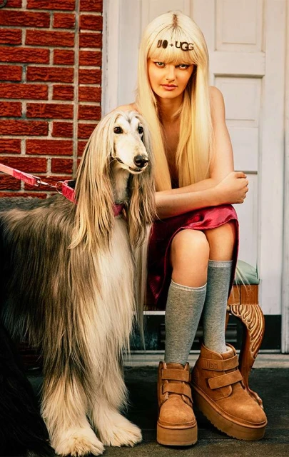 A blonde woman with UGG headband sitting next to an Afghan Hound dog in brown UGG platform boots, against a brick wall and white door.