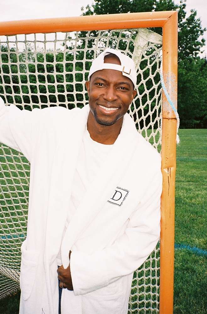 Smiling man in white robe and baseball cap poses with orange lacrosse goal on a green lawn. Trees are visible in the background.