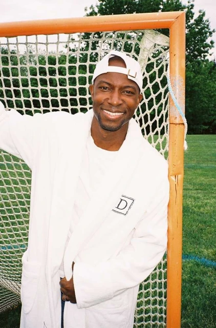 Smiling man in white robe and baseball cap poses with orange lacrosse goal on a green lawn. Trees are visible in the background.
