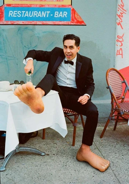 Man in tuxedo opening champagne with feet on table outside restaurant; unique perspective and upscale setting for celebration or lifestyle.