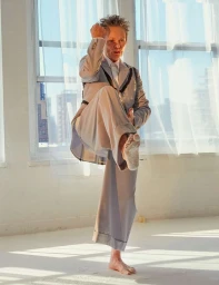 An older woman in a grey suit holding a martial arts pose barefoot before large windows with sheer curtains.