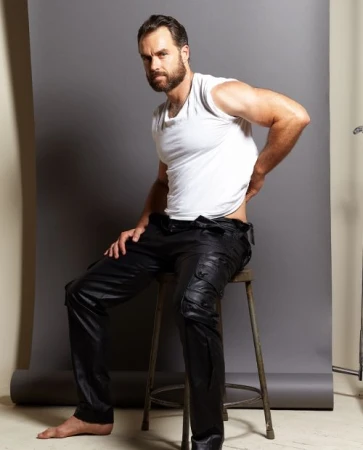Man with beard seated on metal stool wearing white t-shirt and black cargo pants against grey seamless backdrop