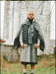 Portrait of a man with a shaved head wearing layered grey and black clothing outdoors with a silo in the background