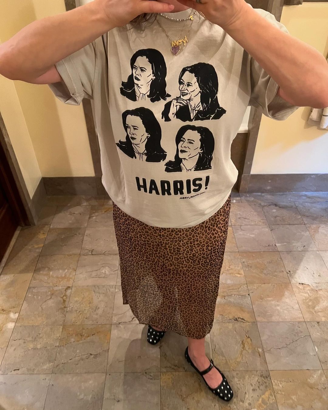 Woman in t-shirt with Kamala Harris portraits and leopard print skirt stands on tiled floor in an eye-level shot.