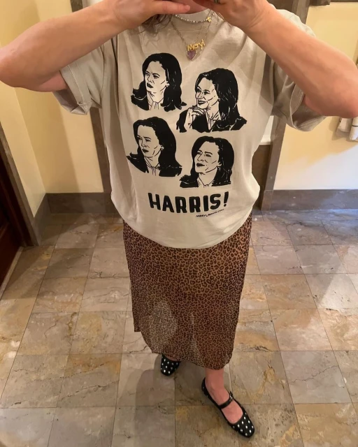 Woman in t-shirt with Kamala Harris portraits and leopard print skirt stands on tiled floor in an eye-level shot.