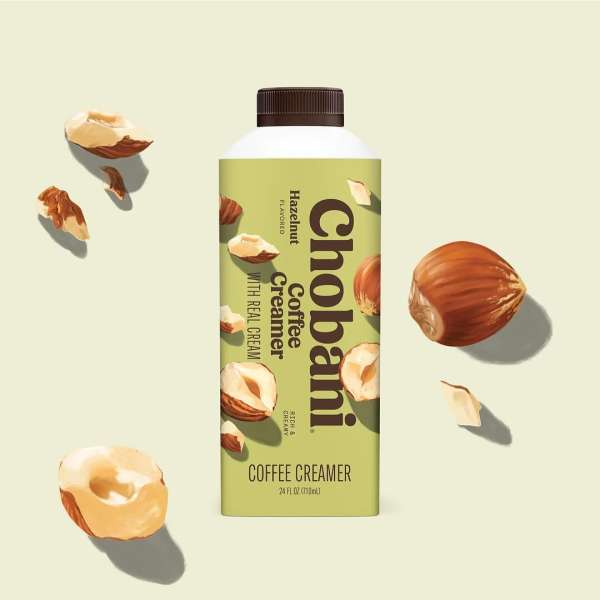 Chobani Hazelnut coffee creamer in a carton with scattered hazelnuts against a cream-colored background in a studio product shot.
