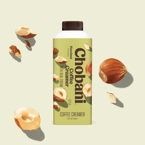 Chobani Hazelnut coffee creamer in a carton with scattered hazelnuts against a cream-colored background in a studio product shot.