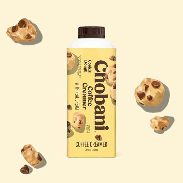 Chobani Cookie Dough Coffee Creamer bottle surrounded by cookie dough pieces against a light yellow background in a studio setting.