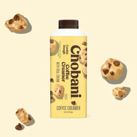 Chobani Cookie Dough Coffee Creamer bottle surrounded by cookie dough pieces against a light yellow background in a studio setting.