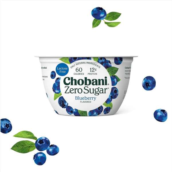 Chobani Zero Sugar Blueberry yogurt container with blueberries and green leaves floating around it on a white background. Food photography.