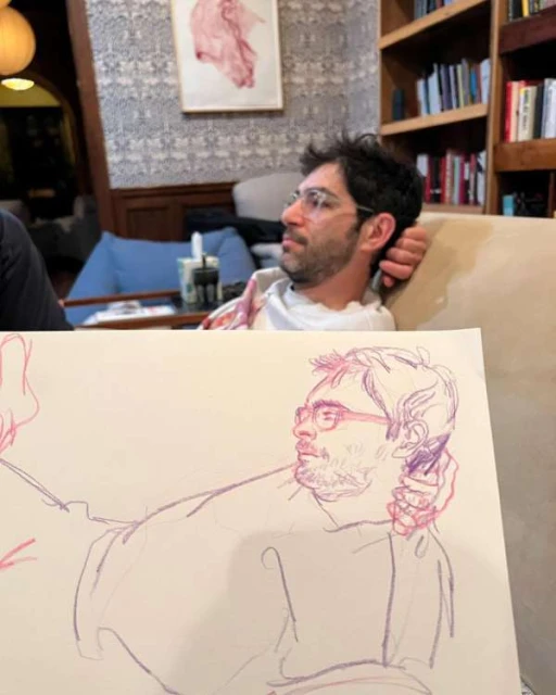 A man with glasses sits on a sofa as an artistic pencil sketch of him is partially visible in the foreground of the image.