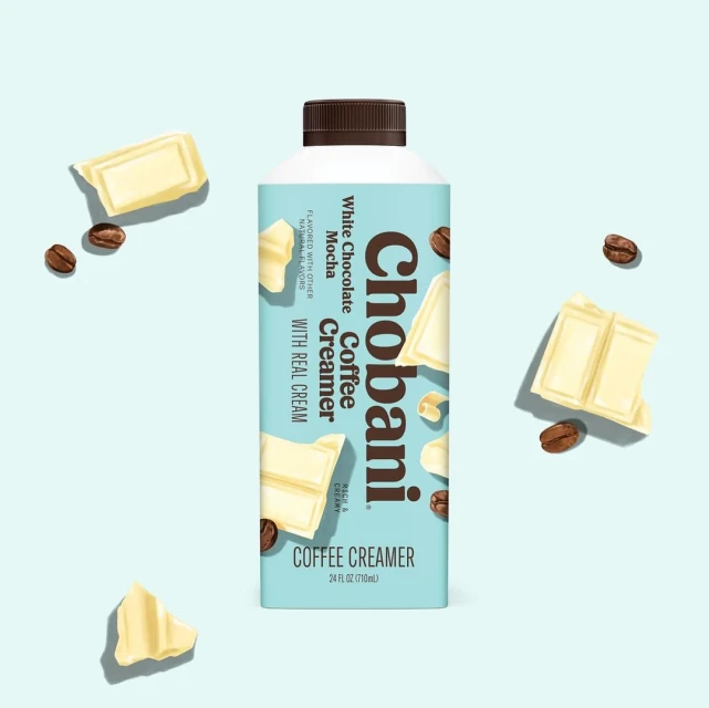 Chobani White Chocolate Mocha Coffee Creamer bottle on a light blue background with scattered white chocolate pieces and coffee beans.