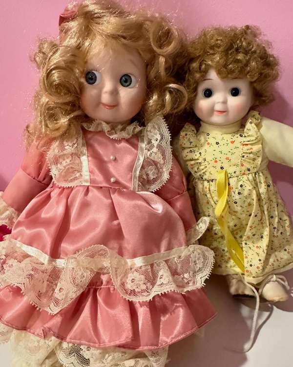 Two vintage porcelain dolls with curly hair wearing dresses with lace trim on a light pink background, capturing a nostalgic feel.