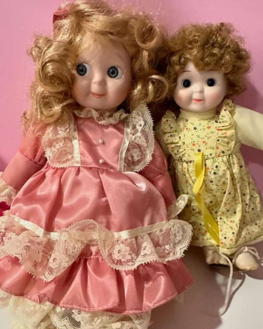Two vintage porcelain dolls with curly hair wearing dresses with lace trim on a light pink background, capturing a nostalgic feel.