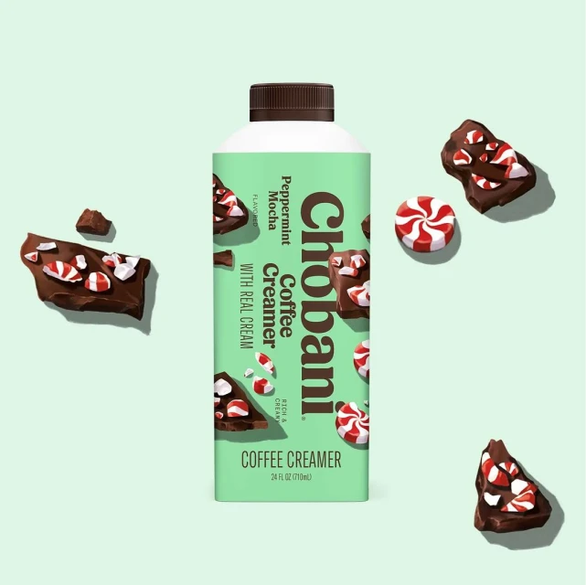 A bottle of Chobani Peppermint Mocha coffee creamer is staged with chocolate peppermint bark on a light green backdrop.