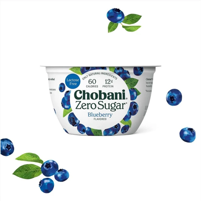 Chobani Zero Sugar Blueberry yogurt in a white container surrounded by blueberries and leaves against a white background.