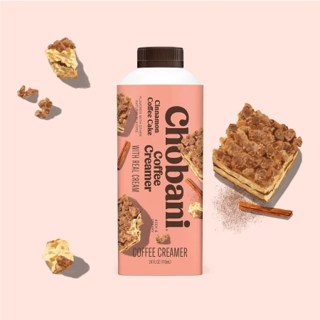 Chobani Cinnamon Coffee Cake Creamer bottle, cake pieces, and cinnamon sticks on a light pink backdrop in a studio shot.