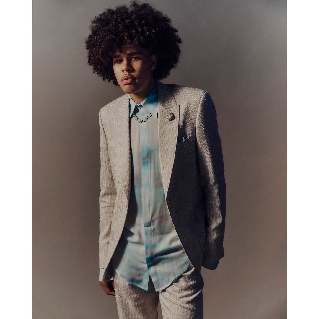 Young adult male with afro in a light beige suit and patterned shirt in a portrait shot against a gray studio backdrop.