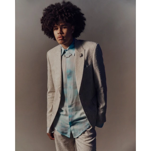 Young adult male with afro in a light beige suit and patterned shirt in a portrait shot against a gray studio backdrop.