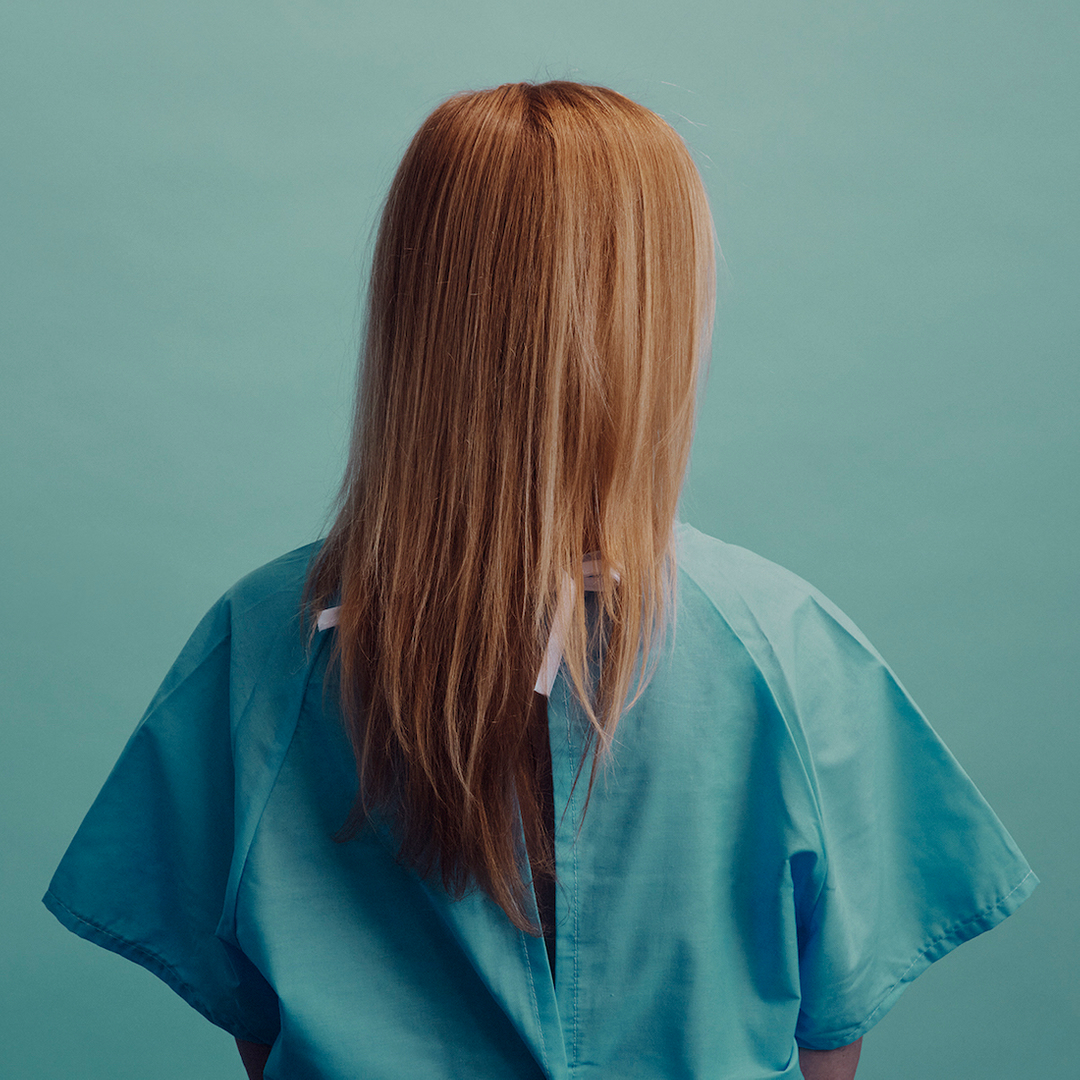 Back view of person with strawberry blonde hair wearing a blue surgical gown against an aqua background in a studio setting.