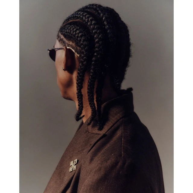 Portrait of an adult male with braided hair and sunglasses, wearing a brown blazer with a square brooch, against a neutral gray backdrop.