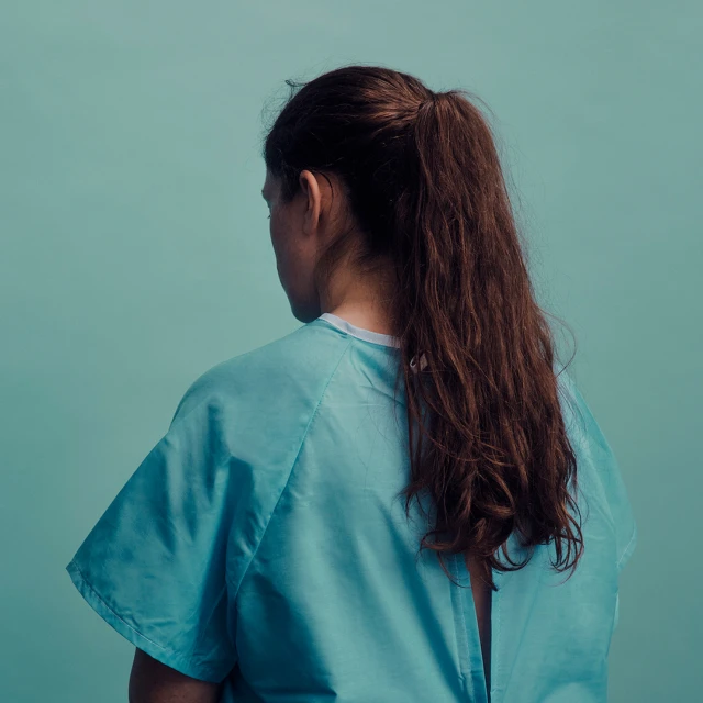 Back view of a woman with long brown hair in a high ponytail, wearing a blue medical gown, standing in front of a light blue background.