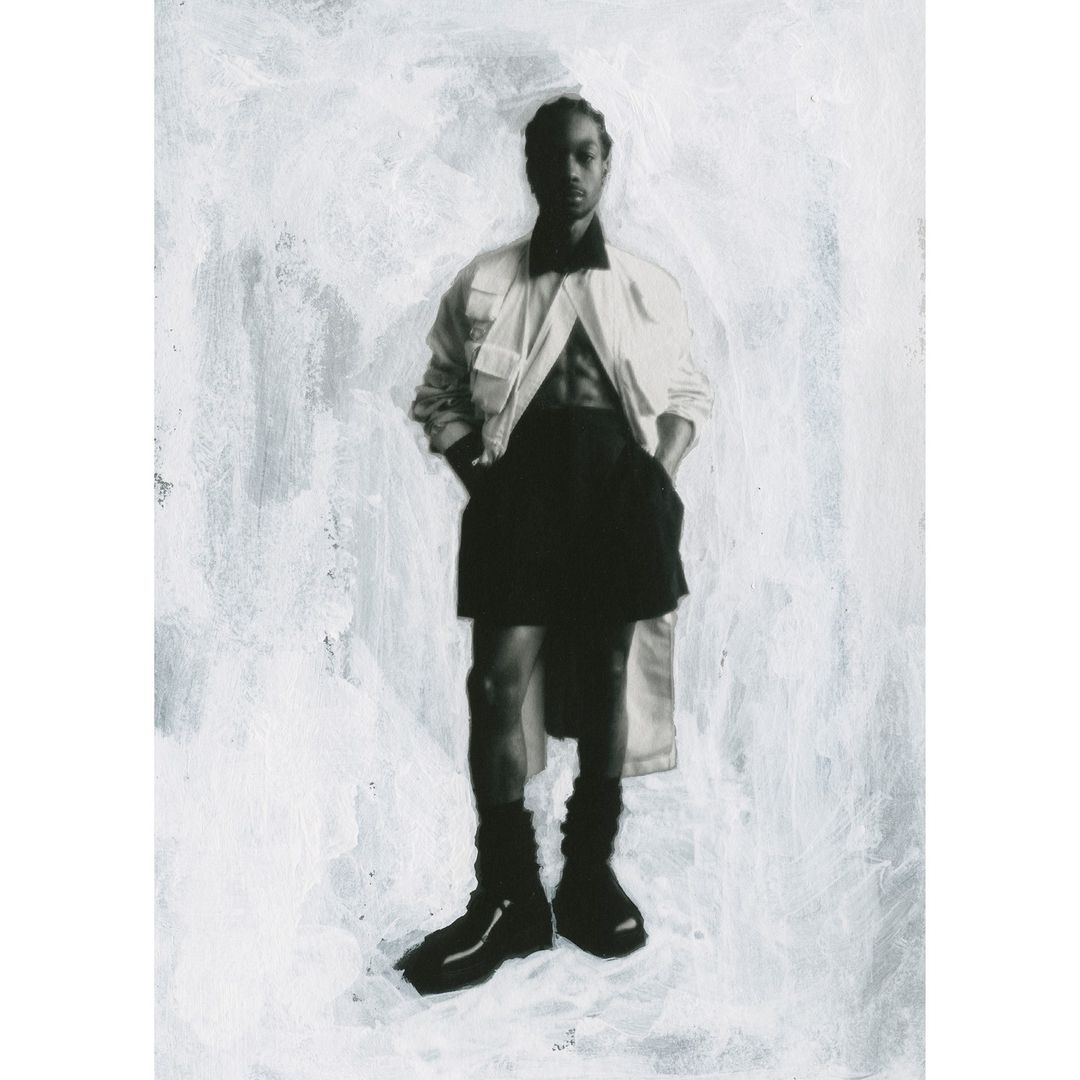 Fashionable person in a white jacket, black skirt, and boots against a textured white background, posing with hands in pockets.