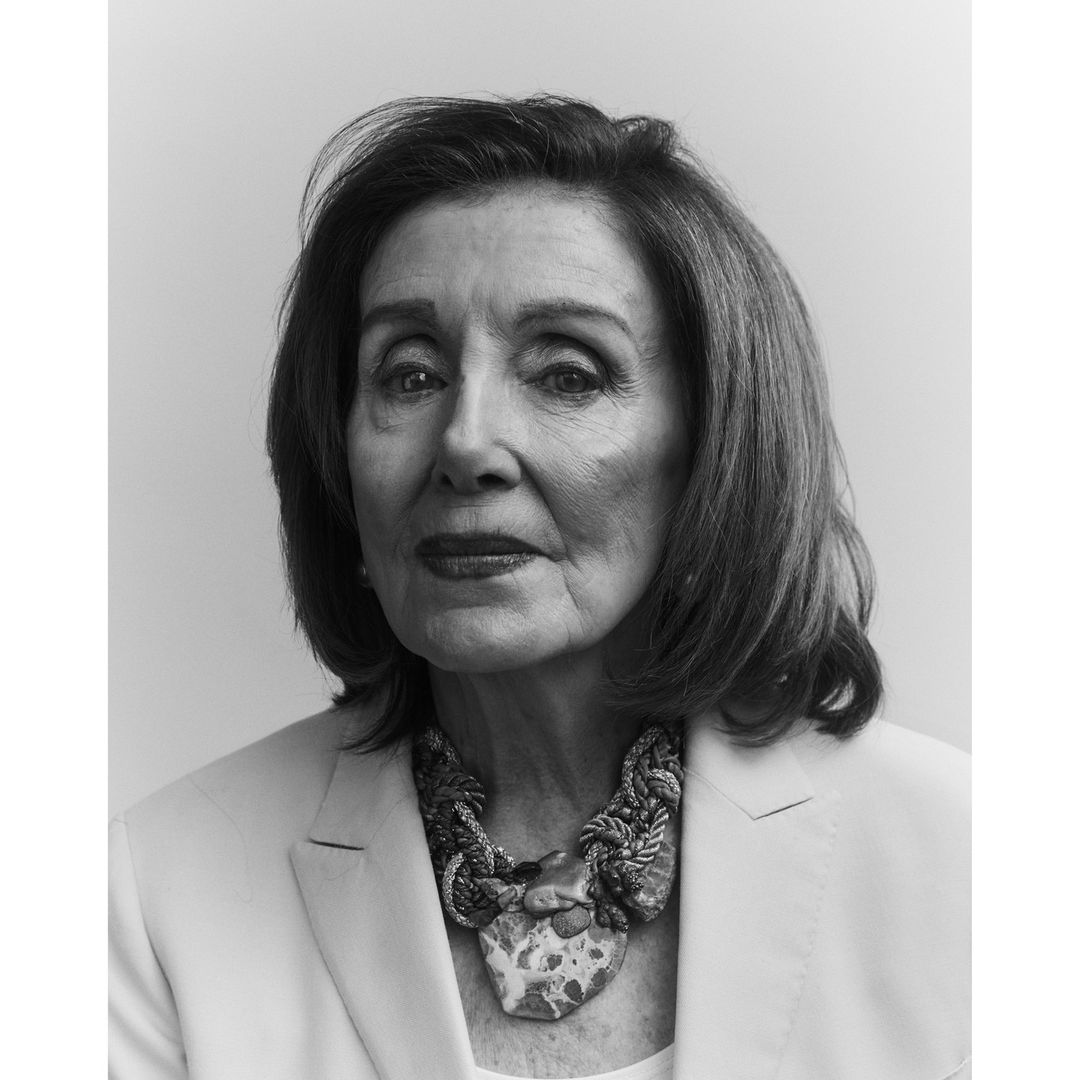 Black and white portrait of an older woman wearing a statement necklace and blazer against a neutral backdrop in a studio setting.