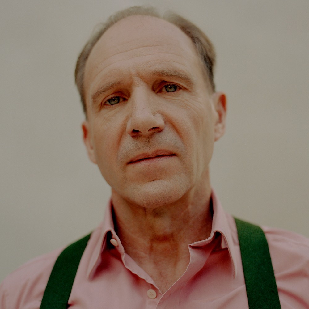 Close-up portrait of an adult male with short hair and wearing a pink collared shirt with green suspenders against a neutral backdrop.