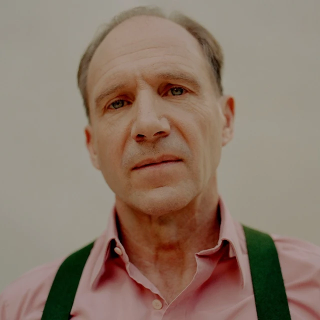 Close-up portrait of an adult male with short hair and wearing a pink collared shirt with green suspenders against a neutral backdrop.