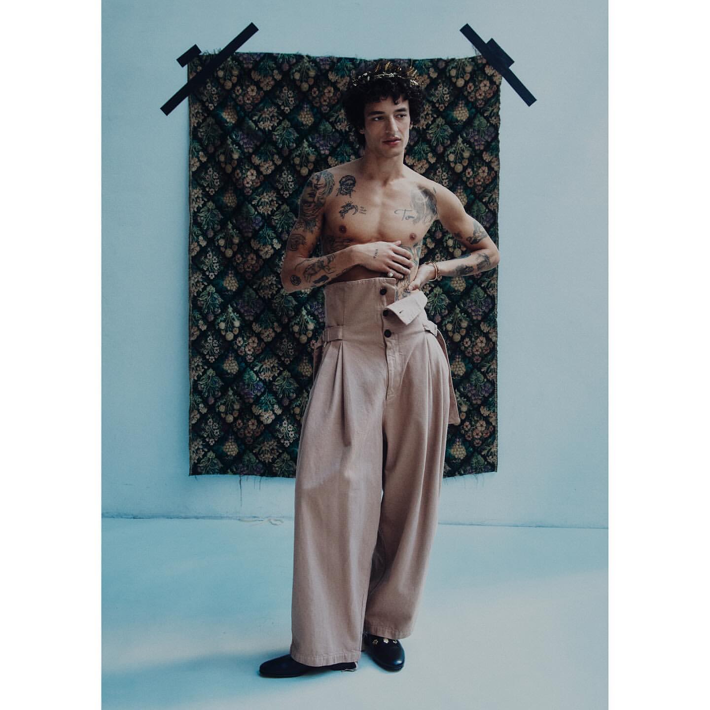 Man with tattoos poses in high-waisted beige trousers against a patterned tapestry in a full shot in a photo studio setting.