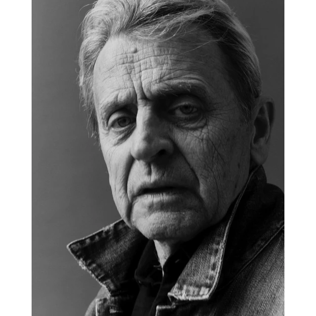 Monochrome portrait of a mature adult male with short grey hair wearing a denim jacket, set against a solid dark grey background.