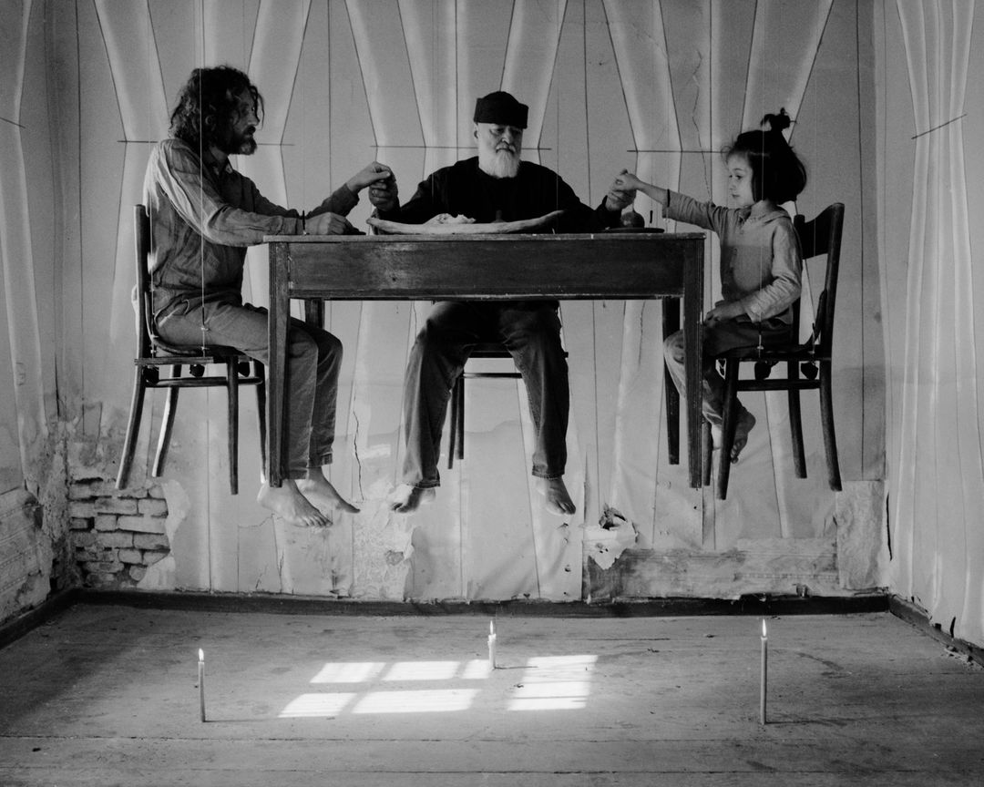 Monochrome image features an adult male, senior adult, and a child at a table, seemingly floating above the floor with lit candles.