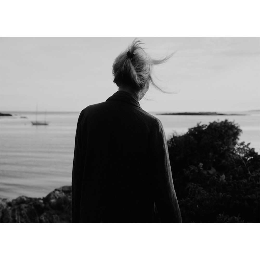 Woman stands on a rocky outcrop overlooking the water, with a sailboat in the distance, set against a monochrome sky.