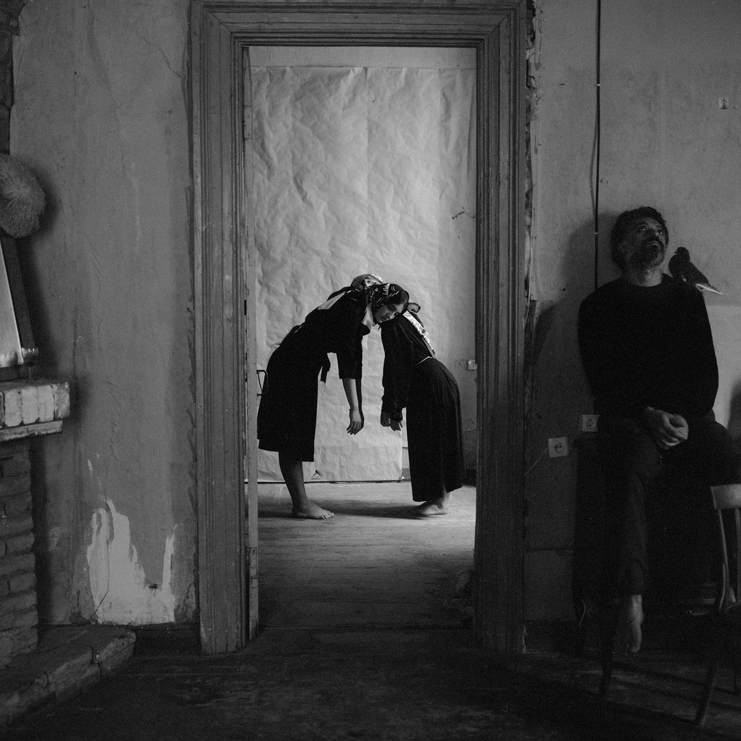Black and white portrait of two women with joined foreheads in black dresses, framed by a doorway, with a man seated on a stool.