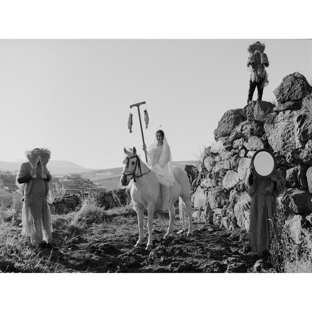 Monochrome image featuring a woman in white on horseback with fish, flanked by two figures and another on a stone wall. Rural landscape.