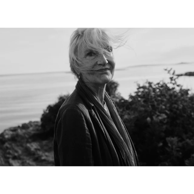 Black-and-white portrait of a mature woman with windswept hair, wearing a dark jacket, with a coastal background on a sunny day.
