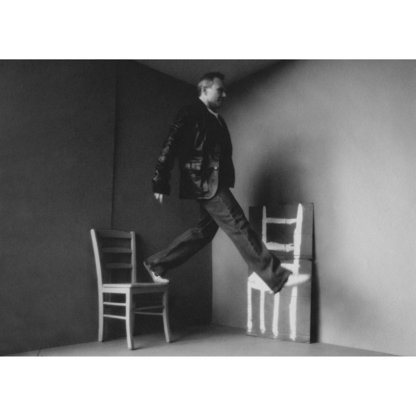Monochrome image of a man in a jacket and jeans leaping over chairs in a sparse room with gray walls and neutral lighting.